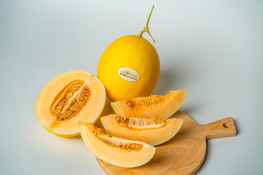 A whole Melonmas melon beside halved and sliced portions on a wooden board, showing the deep orange flesh.